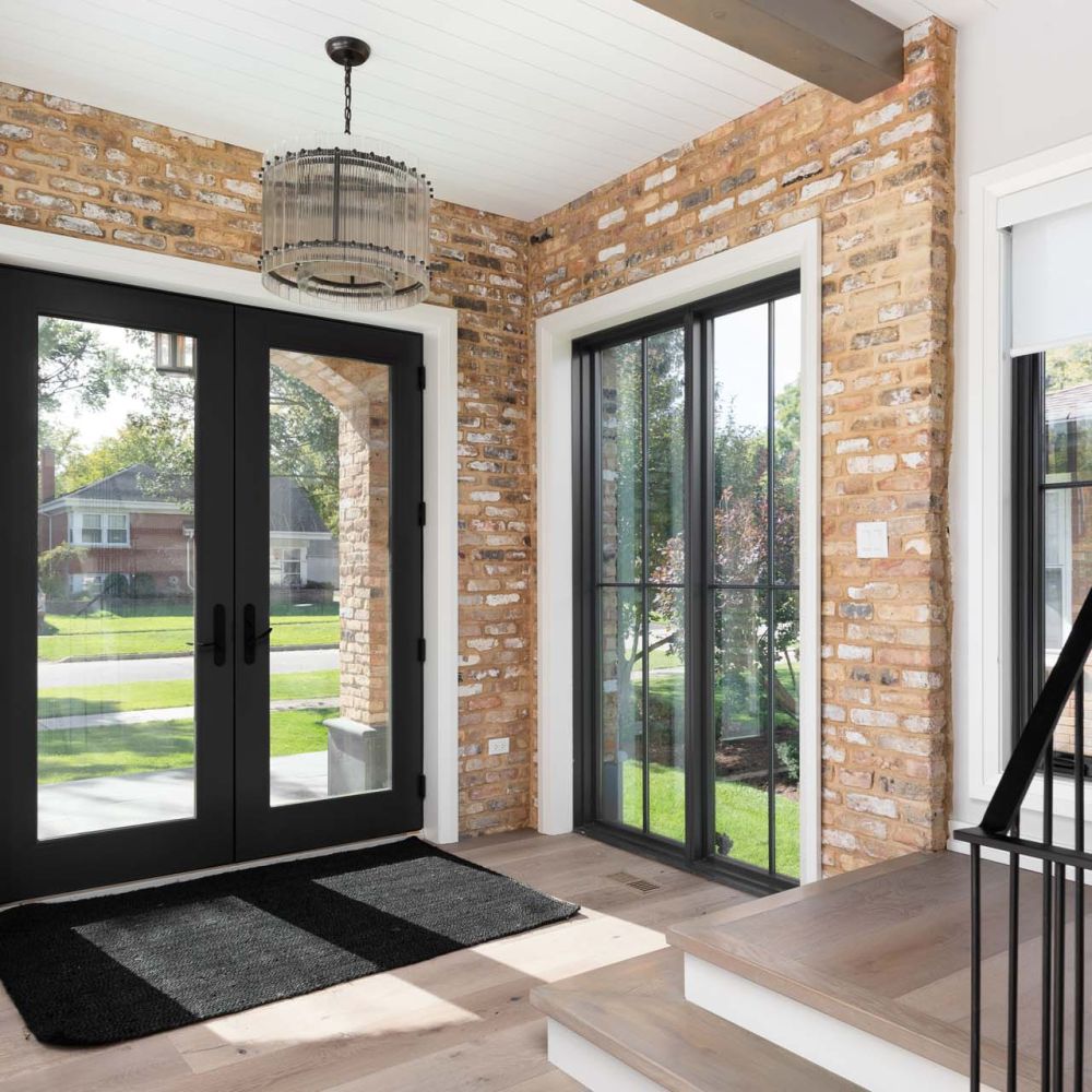 A cozy detail of an entryway with black French doors, brick and white walls, black chandelier hanging from a shiplap ceiling, and hardwood floors and stairs with wrought iron railings.